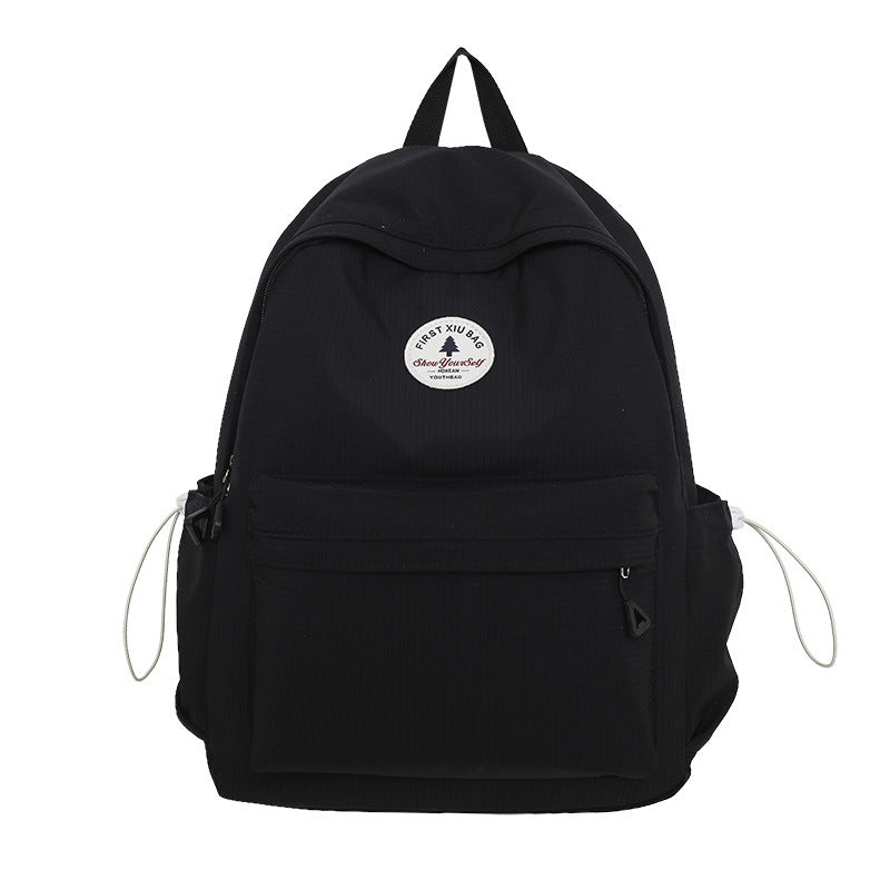 A black nylon backpack with a top handle, a front zippered pocket, side mesh pockets with drawstring cords, and a circular white logo patch on the front.
