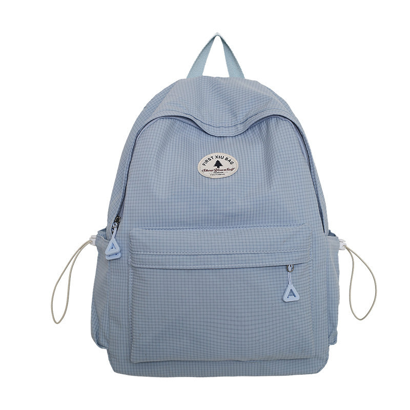 A light blue gingham backpack with a front zippered pocket, side pockets, and a circular 'The Minimalist' brand patch on the front, shown against a plain white background.